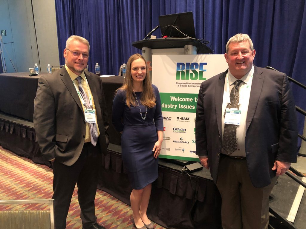 Two men and one woman pose in business attire by a sign with the RISE logo.