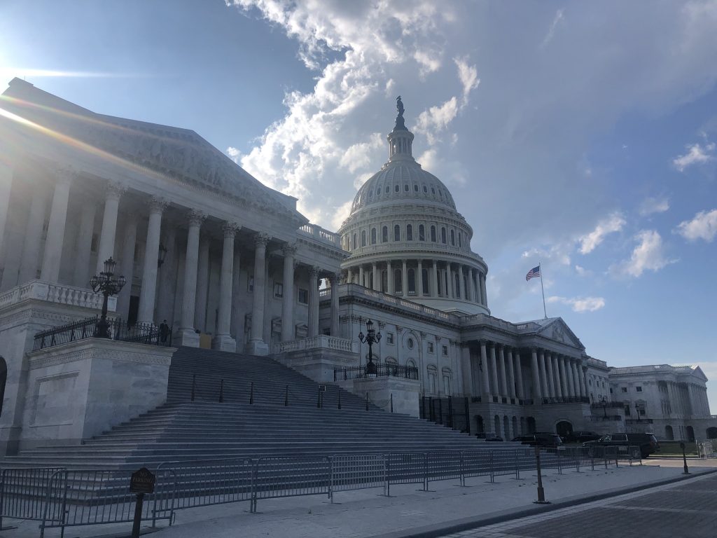 The United States Capitol building with the sun shining behind it and blue skies above. An American flag waves in front.